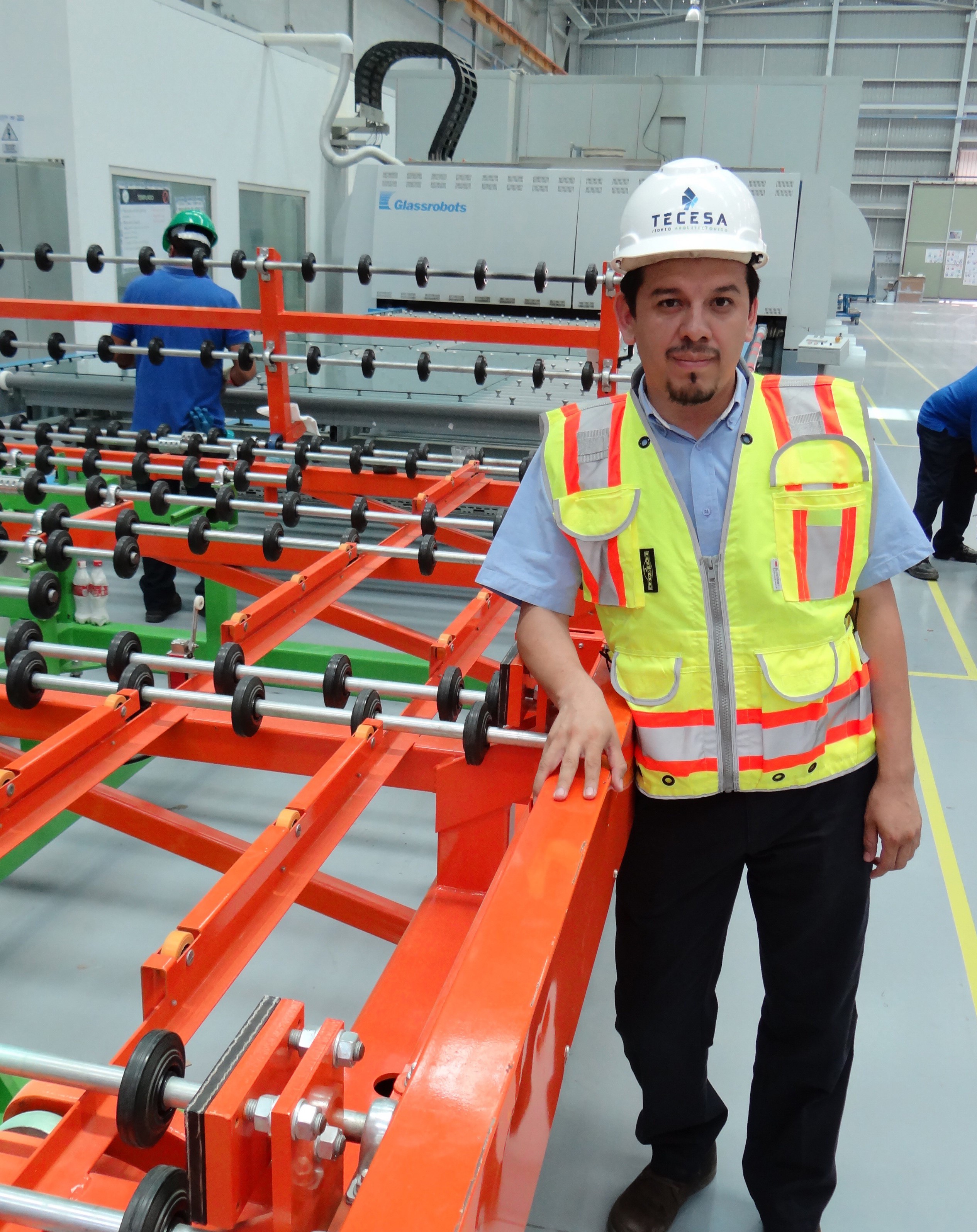 A Tecesa Architectural Glass employee wearing a white hard hat and a high-visibility vest stands next to a workstation featuring a computer monitor displaying FeneVision software. The background shows an industrial glass manufacturing facility.