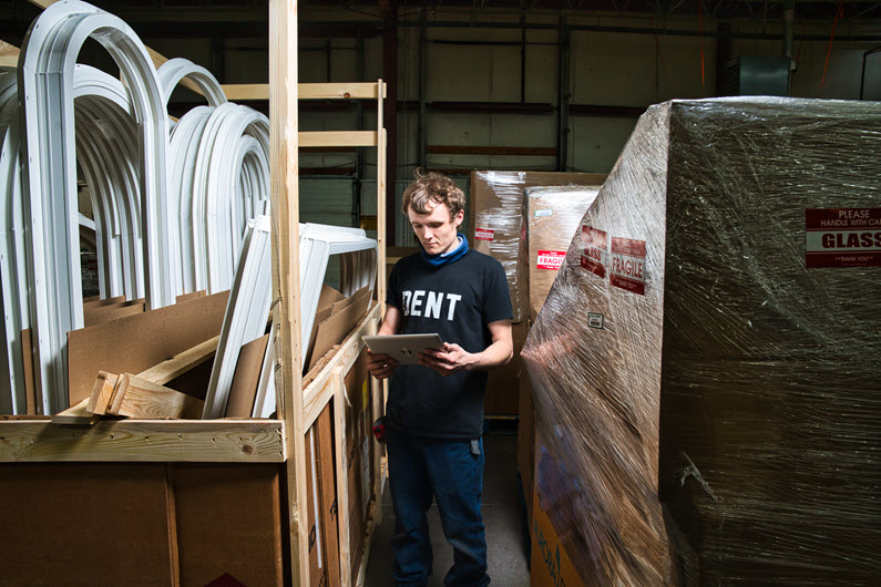 A Fen-Tech worker wearing a 'DENT' T-shirt and a neck gaiter, standing in a storage area filled with various window frames and packaged materials. The worker is holding a tablet and checking inventory.