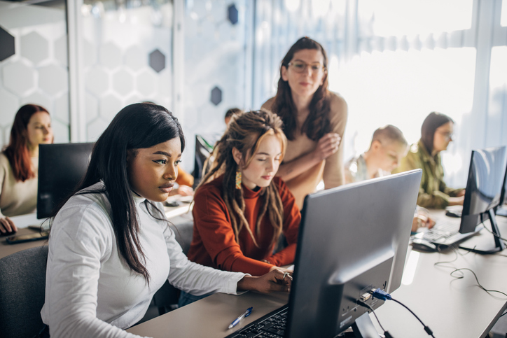 A group of diverse individuals is engaged in a training session in a computer lab. A young woman in a white shirt is focused on her desktop computer, while another woman in a red sweater with long dreadlocks is sitting next to her. An instructor stands nearby, offering guidance. The room has a modern design with hexagonal wall patterns.