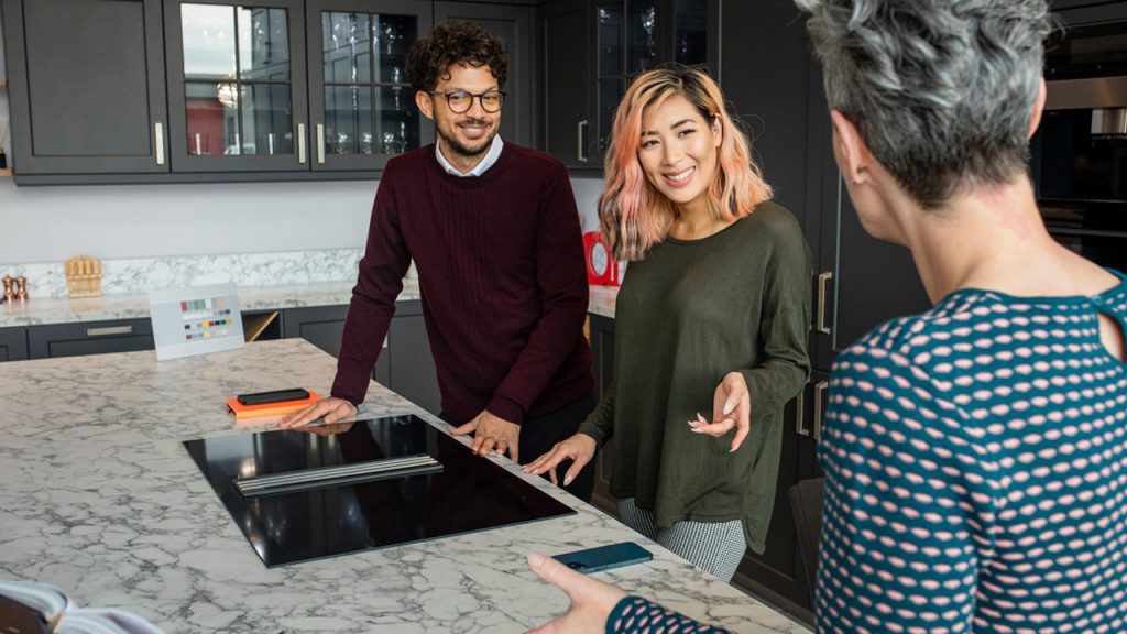 A couple and a woman standing in a kitchen, engaged in conversation.