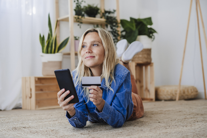 A woman lying on her stomach on the floor, holding a credit card and using a smartphone. The room is decorated with plants and wooden furniture.