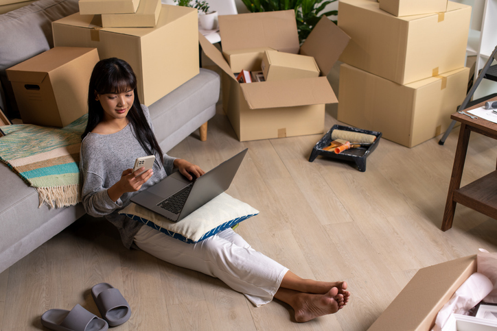 A woman sitting on the floor next to a couch, holding a smartphone and using a laptop. The room is filled with moving boxes, and various personal items are scattered around.