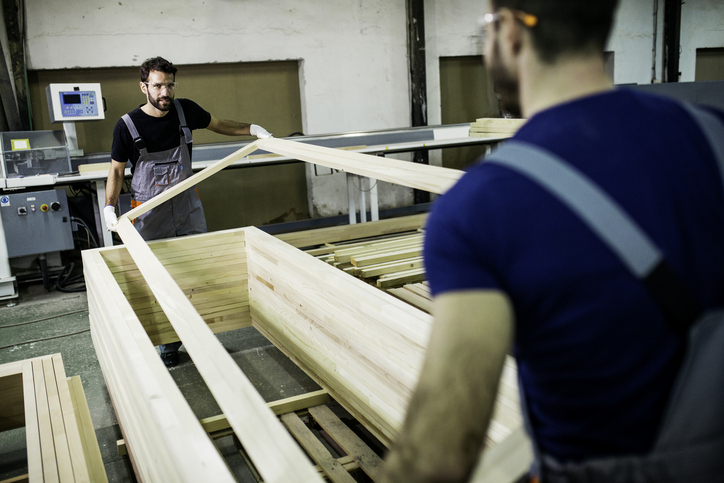 Two workers in a woodworking shop assembling a large wooden frame, wearing safety gear and handling the materials carefully.