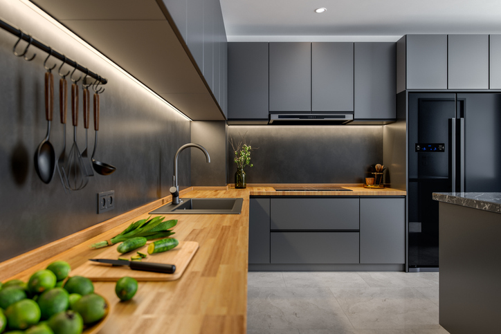 A modern, minimalist kitchen with sleek gray cabinets and a wooden countertop. The kitchen is well-organized with hanging utensils, a built-in sink, and a well-lit workspace. Fresh vegetables are placed on the countertop.