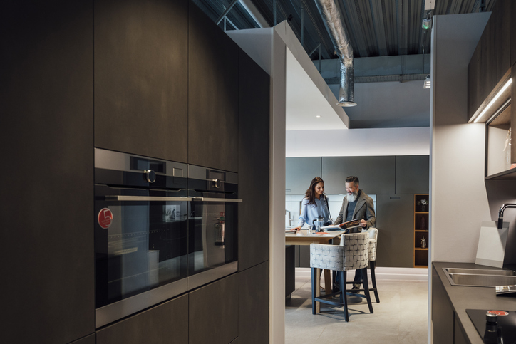 A couple reviews design plans in a modern kitchen showroom. They are seated at a table with samples and a layout plan, with stylish cabinetry and kitchen appliances surrounding them.