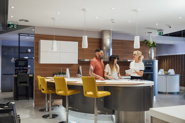 A couple consults with a kitchen designer in a showroom. They are standing around a large island with yellow bar stools, discussing design options with the designer, who is holding a tablet.