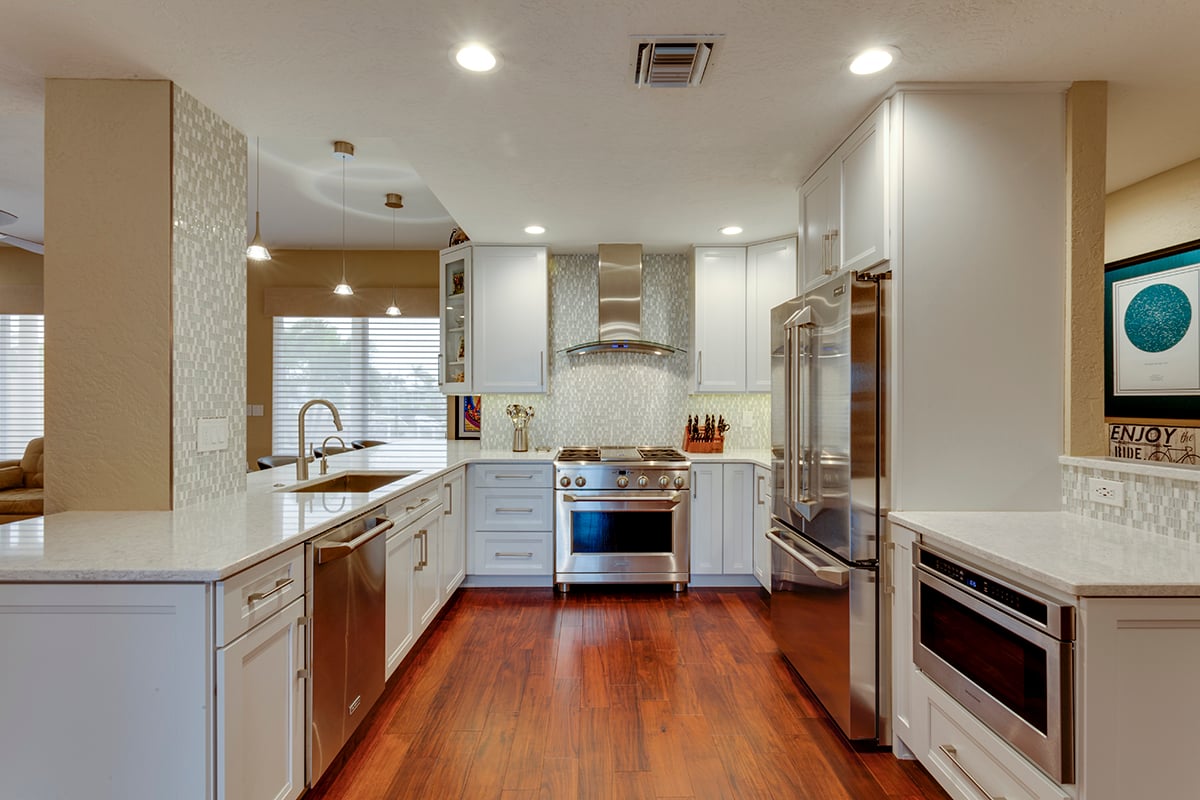 Modern white kitchen with stainless steel appliances, a central island featuring a built-in sink and seating area, glossy white cabinetry, and mosaic backsplash tiling under soft pendant lights.