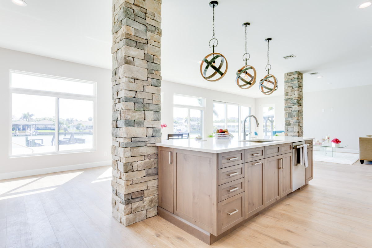 Bright, open-concept kitchen with a central island made of natural wood, stone pillar accents, industrial pendant lighting, and expansive windows overlooking a waterfront view.