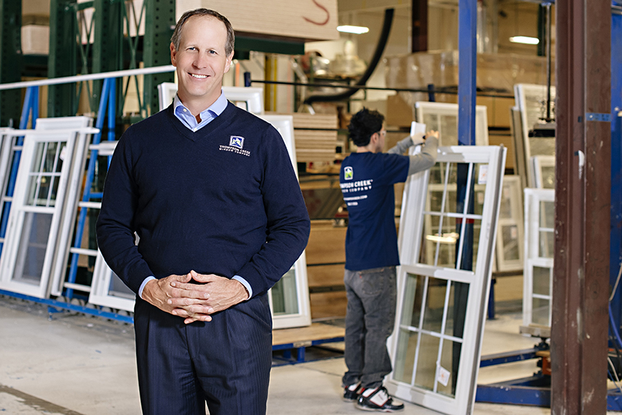 A Thompson Creek representative wearing a navy blue sweater with the company logo stands in front of window frames stacked on racks. A worker in the background assembles a window in the production facility.