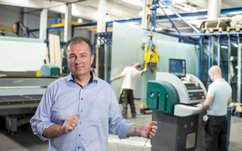 A man stands in front of a factory machine holding a sheet of glass