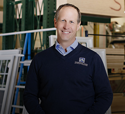 A Thompson Creek representative in a navy blue sweater with the company logo smiles while standing in a window production area, with stacks of frames and a worker assembling windows in the background.